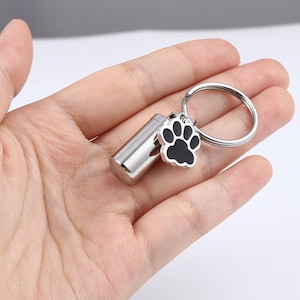 May include: Silver keychain with a small cylindrical container and a black paw print charm. The keychain is designed to hold a small amount of ashes or other mementos.
