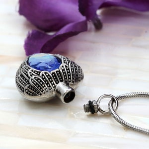May include: Silver heart-shaped cremation pendant with a blue crystal and angel wings. The pendant is open and the chain is visible.