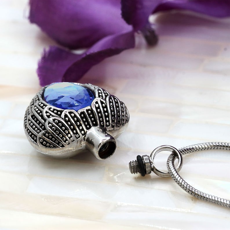 May include: Silver heart-shaped cremation pendant with a blue crystal and angel wings. The pendant is open and the chain is visible.