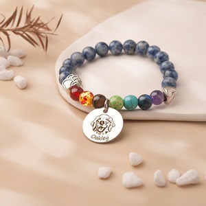 May include: A blue gemstone bracelet with a charm featuring a dog's portrait and the name "Oakley". The charm is attached to a rainbow of smaller gemstones.