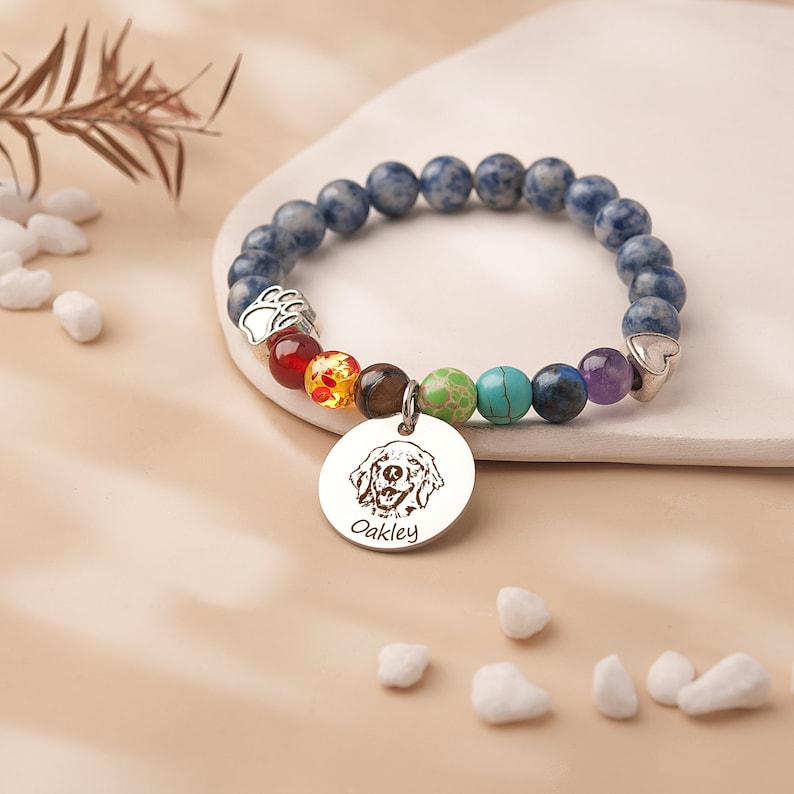 May include: A blue gemstone bracelet with a charm featuring a dog's portrait and the name "Oakley". The charm is attached to a rainbow of smaller gemstones.
