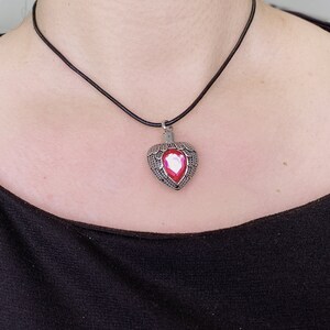 May include: A silver heart-shaped pendant with a pink gem in the center, hanging from a black leather cord necklace.