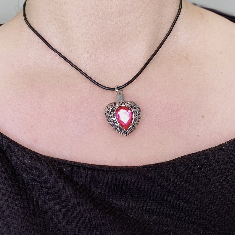 May include: A silver heart-shaped pendant with a pink gem in the center, hanging from a black leather cord necklace.