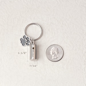 May include: Silver keychain with a small, cylindrical cremation urn, a paw print charm, and the inscription "Mike". The urn is 7/16 inches in diameter and 1 1/8 inches tall. A US quarter is shown for size comparison.