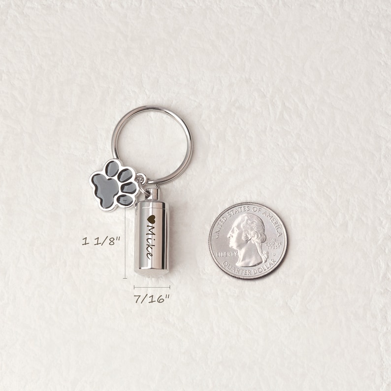 May include: Silver keychain with a small, cylindrical cremation urn, a paw print charm, and the inscription "Mike". The urn is 7/16 inches in diameter and 1 1/8 inches tall. A US quarter is shown for size comparison.