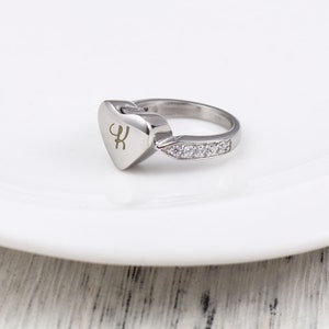 May include: A silver heart-shaped ring with a small inscription and a row of clear cubic zirconia stones on the band.
