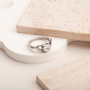 May include: A silver ring with a heart-shaped design. The heart has a floral engraving. The ring is on a white surface.
