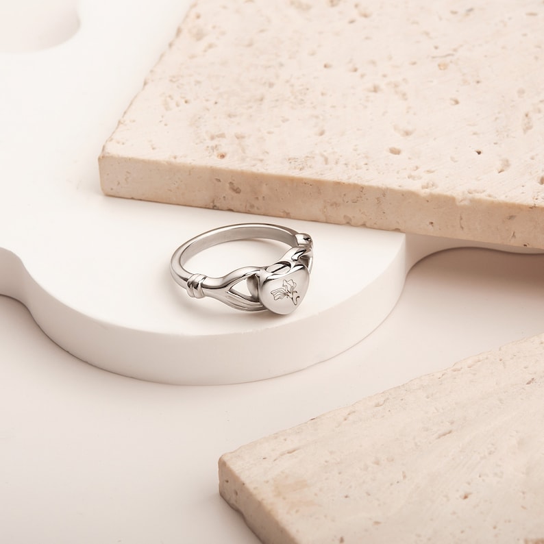 May include: A silver ring with a heart-shaped design. The heart has a floral engraving. The ring is on a white surface.
