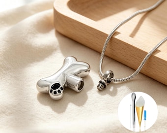 May include: A silver-toned metal cremation pendant shaped like a dog bone with a black paw print detail. The pendant features a threaded screw top and is attached to a silver snake chain. A small circular inset shows a metal funnel and screwdriver.