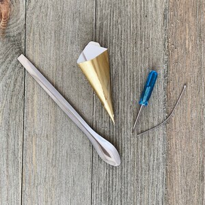 Puede incluir: Un cono de papel dorado y blanco, una cuchara de metal plateado, un destornillador azul y un alambre plateado sobre un fondo de madera.