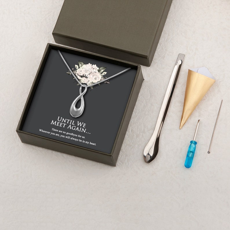 May include: A silver infinity necklace with a white flower bouquet design on a black background. The necklace comes in a black box with the words "Until We Meet Again... There are no goodbyes for us. Wherever you are, you will always be in my heart." The box also includes a small gold cone, a silver tool, a blue screwdriver, and a small silver bead.