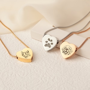 May include: Three heart-shaped cremation necklaces in gold, silver and rose gold, each with an engraved pet portrait. The silver necklace features a paw print, the gold necklace features a cat, and the rose gold necklace features a dog.