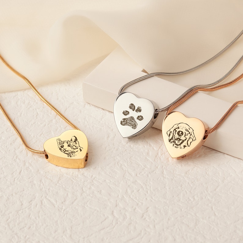 May include: Three heart-shaped cremation necklaces in gold, silver and rose gold, each with an engraved pet portrait. The silver necklace features a paw print, the gold necklace features a cat, and the rose gold necklace features a dog.