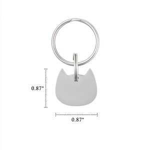 Puede incluir: Una placa de metal plateado con forma de cabeza de gato unida a una anilla circular. La placa tiene una superficie lisa y en blanco, y mide 2,21 cm de alto por 2,21 cm de ancho, tal como indican las l&iacute;neas de dimensi&oacute;n visibles en la imagen.