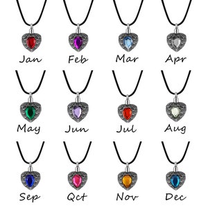 May include: Twelve silver heart-shaped cremation pendants with different colored gemstones. Each pendant is on a black cord and labeled with a month of the year, from January to December.