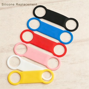 May include: A set of six silicone replacement bands in black, blue, red, pink, white, and yellow. Each band features a central rectangular panel with a circular opening at each end. The text "Silicone Replacement" is printed in the top left corner.