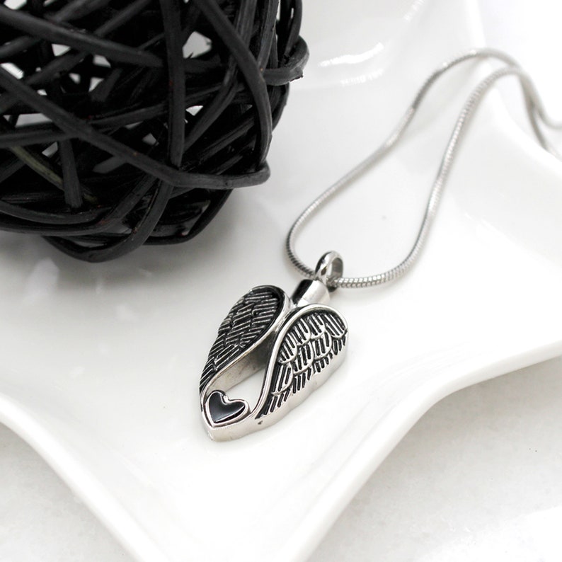 Puede incluir: Un collar de plata con un colgante en forma de alas de &aacute;ngel con un coraz&oacute;n en el centro. El colgante es una urna funeraria.