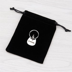 Puede incluir: Una placa de identificaci&oacute;n para mascotas de metal plateado con forma de cabeza de gato y el nombre Luna grabado en la parte frontal, unida a una anilla met&aacute;lica circular, sobre una peque&ntilde;a bolsa de terciopelo negro con cordones.