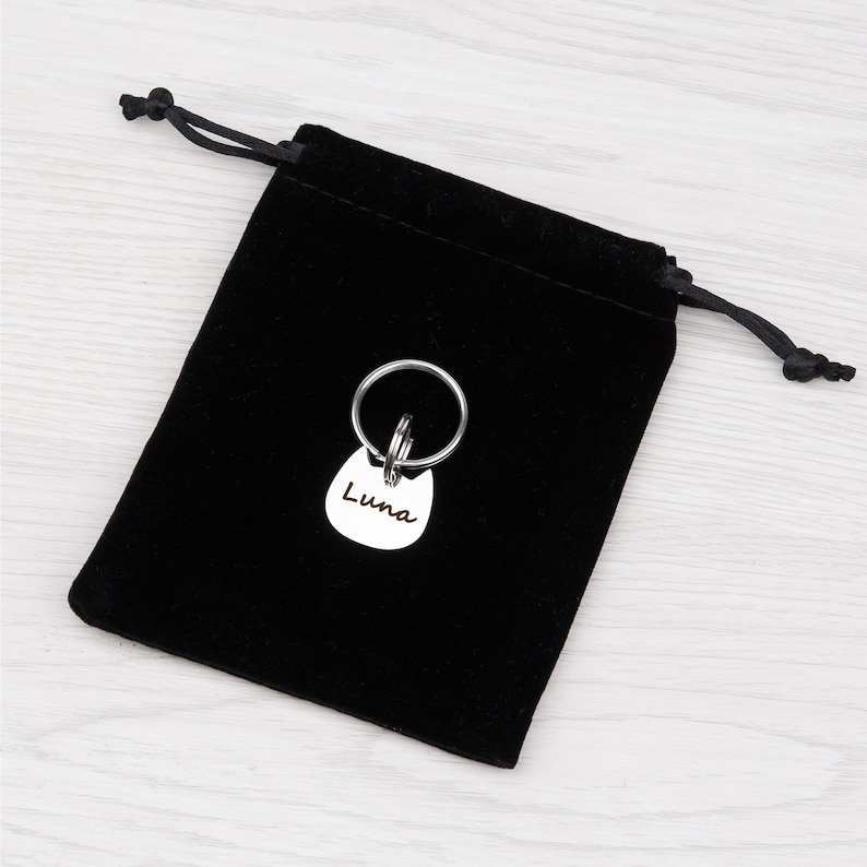 Puede incluir: Una placa de identificaci&oacute;n para mascotas de metal plateado con forma de cabeza de gato y el nombre Luna grabado en la parte frontal, unida a una anilla met&aacute;lica circular, sobre una peque&ntilde;a bolsa de terciopelo negro con cordones.
