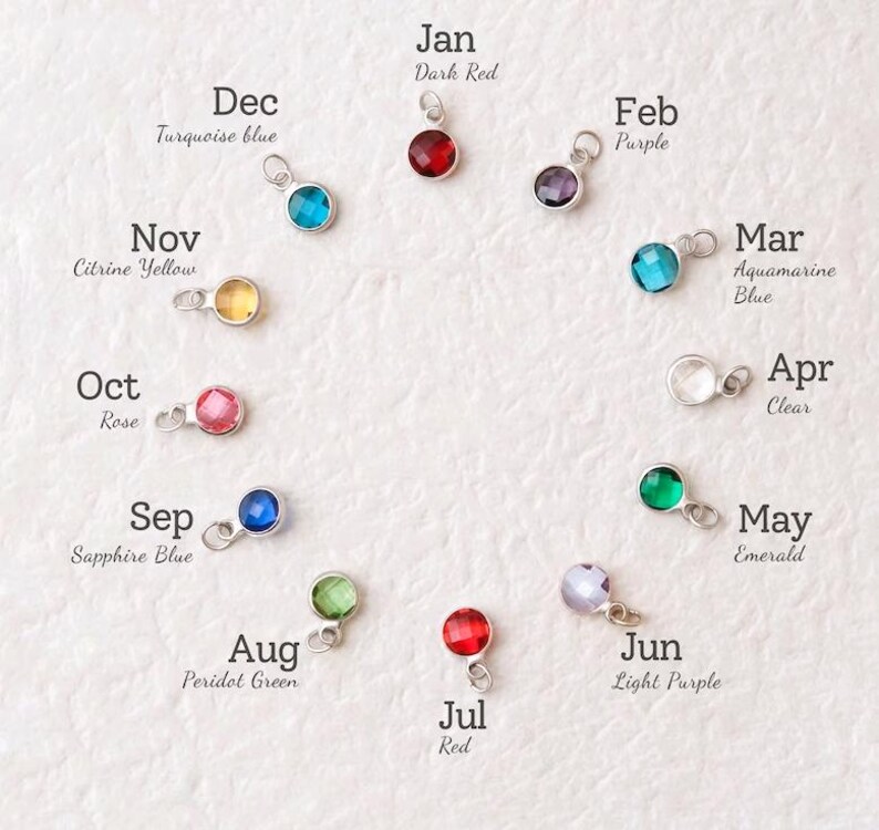 May include: A collection of twelve gemstone charms, each representing a month of the year. The charms are in various colors, including red, purple, blue, green, and yellow, with the month's name and color description next to each charm.
