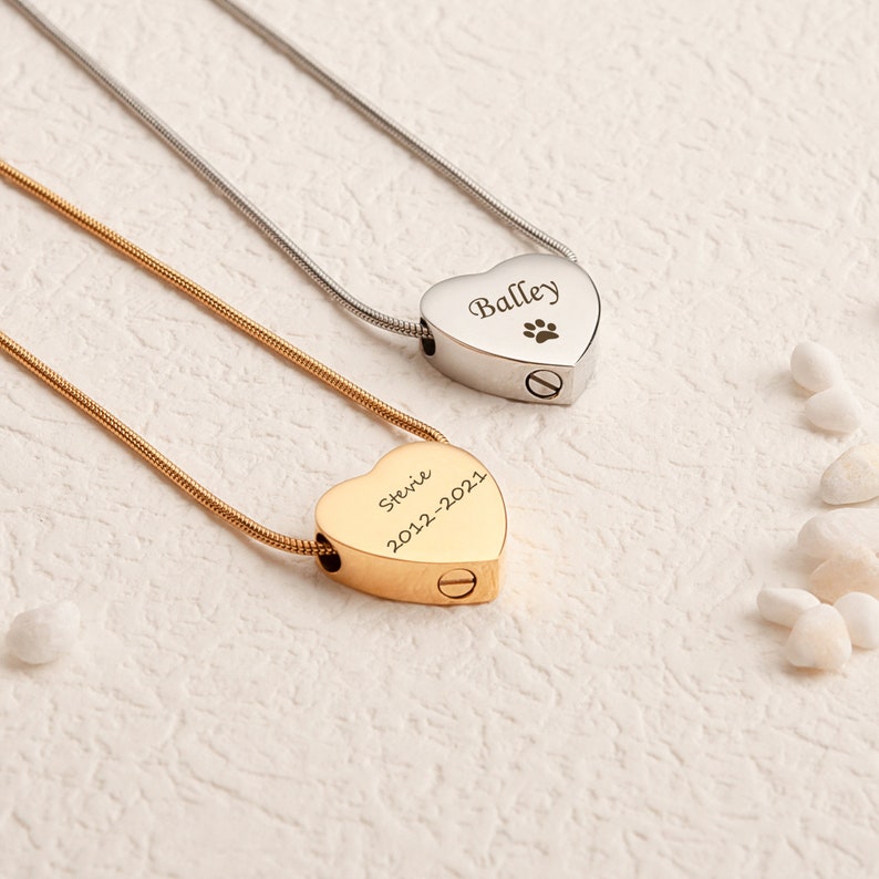 Pu&ograve; includere: Due collane crematorie a forma di cuore, una in oro e una in argento, con nomi e date incisi. La collana d'oro dice "Stevie 2012-2021" e la collana d'argento dice "Bailey" con un'impronta di zampa.