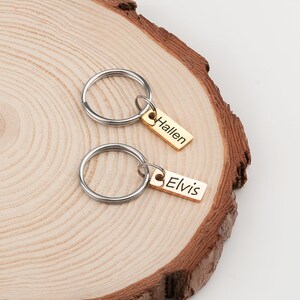 May include: Two gold-toned keyrings with silver key rings. The keyrings have rectangular tags with the names "Elvis" and "Hallen" engraved on them.