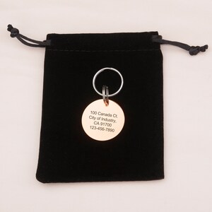 May include: A round, gold-colored metal keychain tag with the text "100 Canada Ct. City of Industry, CA 91700 123-456-7890" engraved on it. The tag is attached to a silver key ring and is sitting on a black velvet pouch.