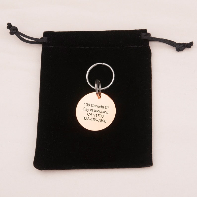May include: A round, gold-colored metal keychain tag with the text "100 Canada Ct. City of Industry, CA 91700 123-456-7890" engraved on it. The tag is attached to a silver key ring and is sitting on a black velvet pouch.
