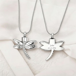May include: Two silver dragonfly pendant necklaces. One pendant features a detailed dragonfly design. The other pendant is engraved with the name "John" and the date "03.21.1996" on the wings.