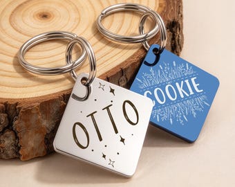 May include: Two square metal pet ID tags with silver-toned split rings. One tag is silver with the name OTTO and small star icons engraved in black. The second tag is blue with the name COOKIE and a white botanical illustration.