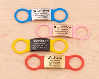May include: Four silicone pet collar tags with metal plates. The blue tag reads Dexter with paw prints, the pink tag reads SNOOPY with hearts, the yellow tag reads Charlotte, and the red tag reads Willow with contact information and microchip status.