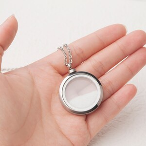 Pode incluir: Um colar de corrente de prata com um pingente redondo de prata. O medalh&atilde;o tem um inserto de vidro transparente e um interior branco.