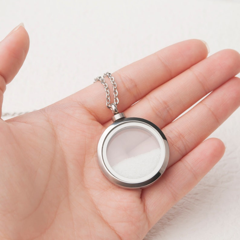 Pode incluir: Um colar de corrente de prata com um pingente redondo de prata. O medalh&atilde;o tem um inserto de vidro transparente e um interior branco.