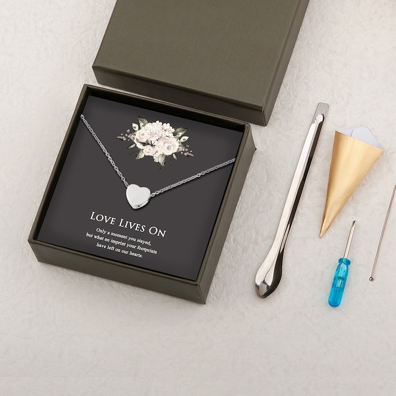 Puede incluir: Un collar de plata con un colgante en forma de coraz&oacute;n en una caja negra con un dise&ntilde;o floral blanco. La caja tiene un mensaje que dice: "El amor sigue vivo. Solo estuviste un momento, pero la huella de tus pasos ha quedado grabada en nuestros corazones."