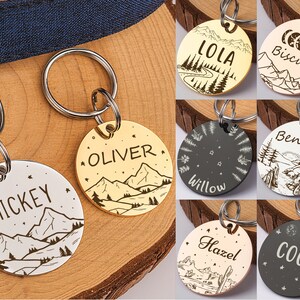 Op de afbeelding: Acht ronde hondenhangers met verschillende ontwerpen en namen. De hangers zijn zilver, goud en zwart met berg-, ster- en maanmotieven. De namen op de hangers zijn Mickey, Oliver, Lola, Biscuit, Willow, Benny, Hazel en Coco.