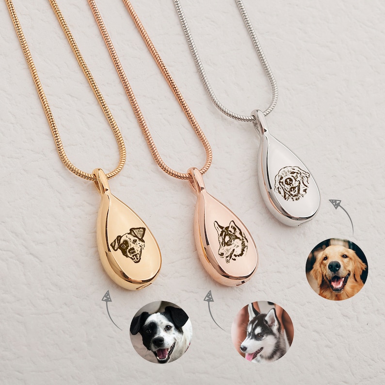 May include: Three silver, gold, and rose gold teardrop-shaped cremation necklaces with engraved images of dogs. The necklaces are on a white background.