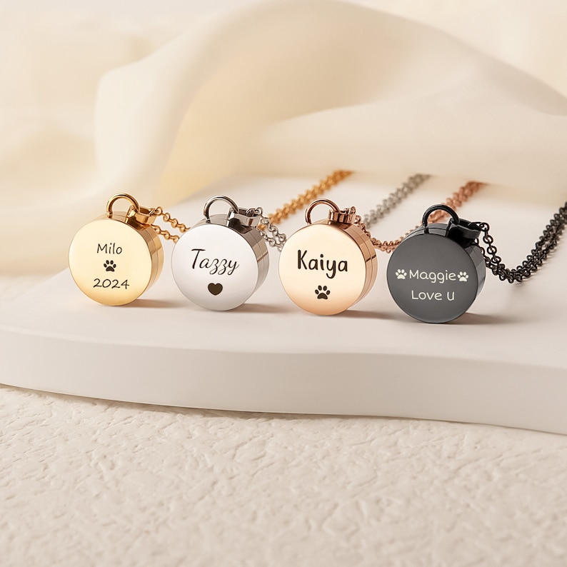 K&ouml;nnte beinhalten: Vier runde Anh&auml;nger in Silber, Gold, Ros&eacute;gold und Schwarz mit gravierten Namen und Pfotenabdr&uuml;cken. Die Anh&auml;nger sind an Ketten und sind ein Denkmal f&uuml;r Haustiere.