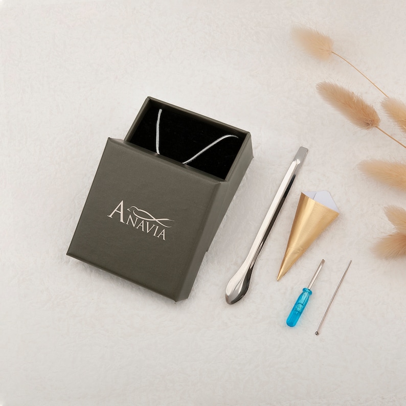 Puede incluir: Un collar plateado en una caja verde oscuro con la palabra "Anavia" impresa en la caja. La caja est&aacute; sobre una superficie blanca con un cono de papel dorado y blanco, una herramienta plateada, un destornillador azul y una aguja plateada.