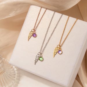 May include: Three necklaces with delicate chains. Each necklace features a wing charm and a gemstone pendant. One necklace is rose gold with a purple gem, another is silver with a green gem, and the third is gold with a purple gem.