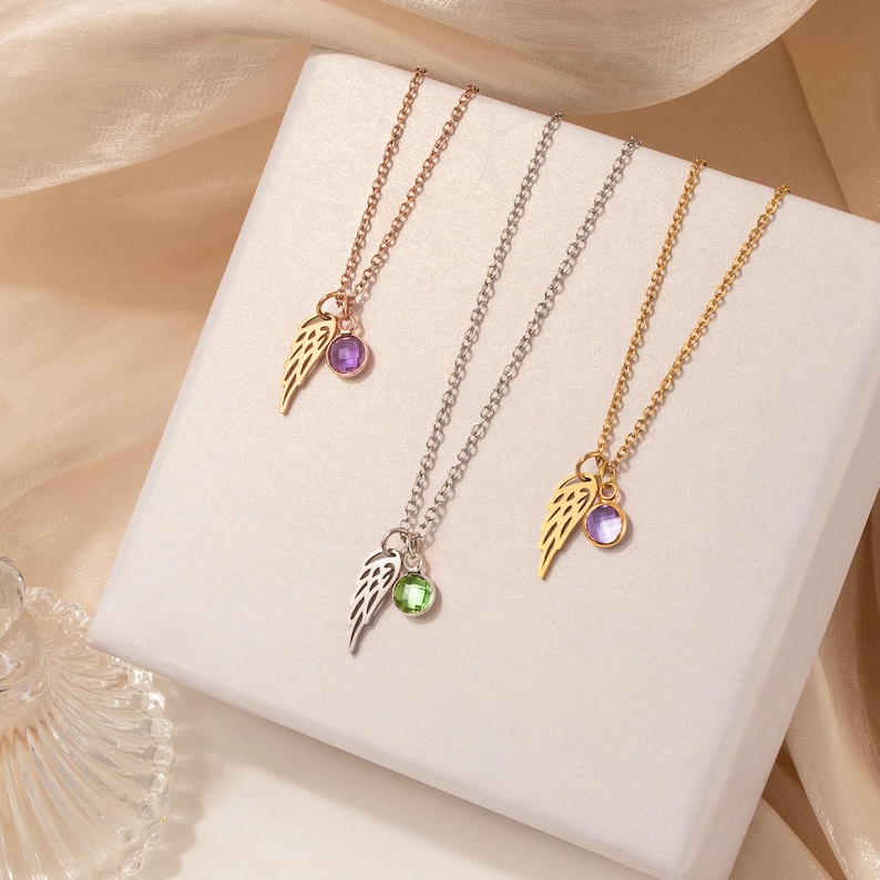 May include: Three necklaces with delicate chains. Each necklace features a wing charm and a gemstone pendant. One necklace is rose gold with a purple gem, another is silver with a green gem, and the third is gold with a purple gem.
