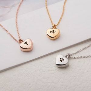 May include: Three gold heart-shaped necklaces with different engravings. The first necklace has a cross engraved on the heart, the second has the name "MAX" engraved on the heart, and the third has a paw print engraved on the heart.
