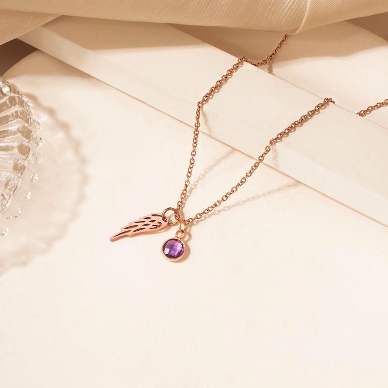 May include: Rose gold necklace featuring a delicate wing charm and a round, purple gemstone pendant. The chain is fine and elegant, with a simple clasp. The necklace is displayed on a neutral background, highlighting its minimalist design.