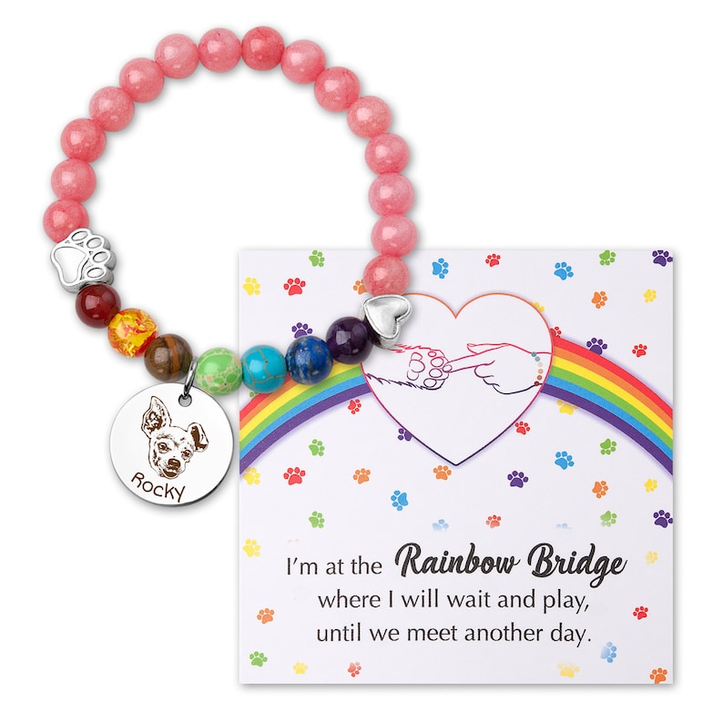 May include: A pink beaded bracelet with a silver charm featuring a paw print and a dog's head. The charm also has the name "Rocky" engraved on it. The bracelet is accompanied by a card with a rainbow and the text "I'm at the Rainbow Bridge where I will wait and play, until we meet another day."