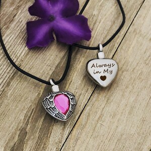 May include: Two silver heart-shaped cremation necklaces. One necklace has a pink crystal in the center of the heart and the other has the words "Always in My" engraved on the front. Both necklaces have black cords.