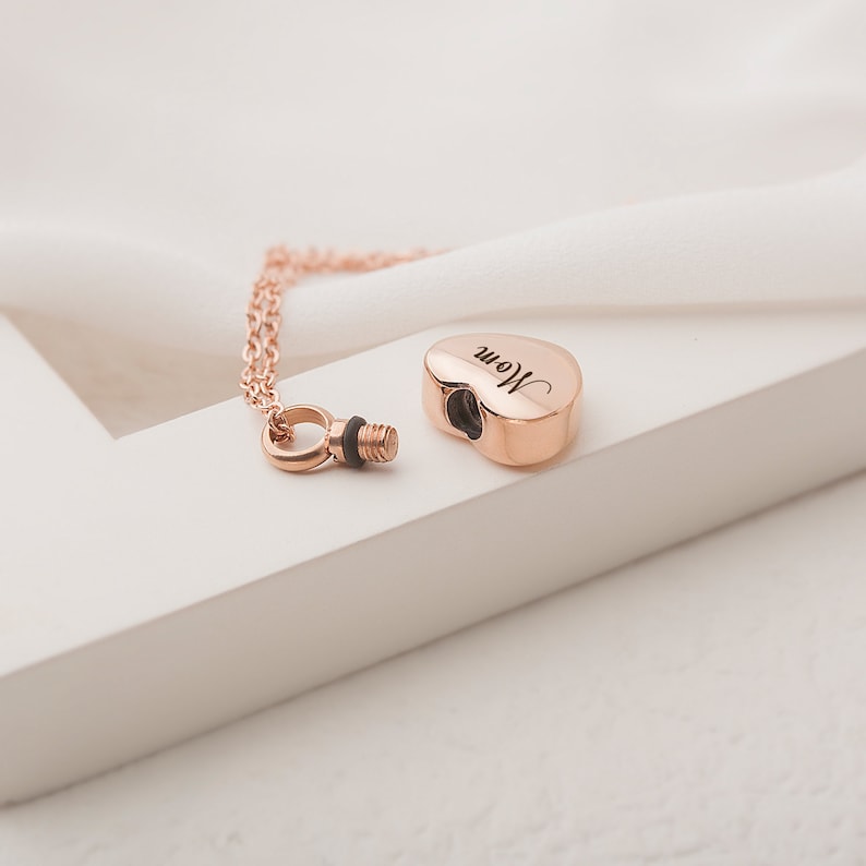 May include: Rose gold heart-shaped cremation necklace with a screw-on lid. The pendant is engraved with the word "Mom".