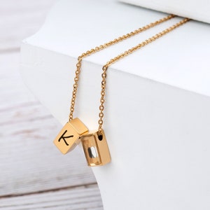 May include: A gold chain necklace with two pendant charms. One charm is a gold cube with the letter "K" engraved on it. The other charm is a clear glass cube with a gold frame.