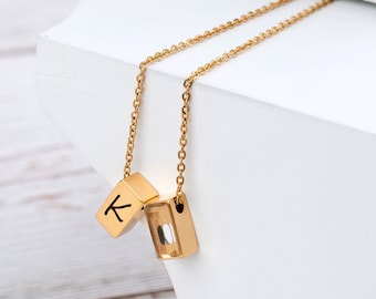 May include: A gold chain necklace with two pendant charms. One charm is a gold cube with the letter "K" engraved on it. The other charm is a clear glass cube with a gold frame.