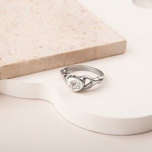 May include: A silver ring with a heart-shaped locket featuring a rose design. The ring is designed to hold a small amount of ashes or other mementos.
