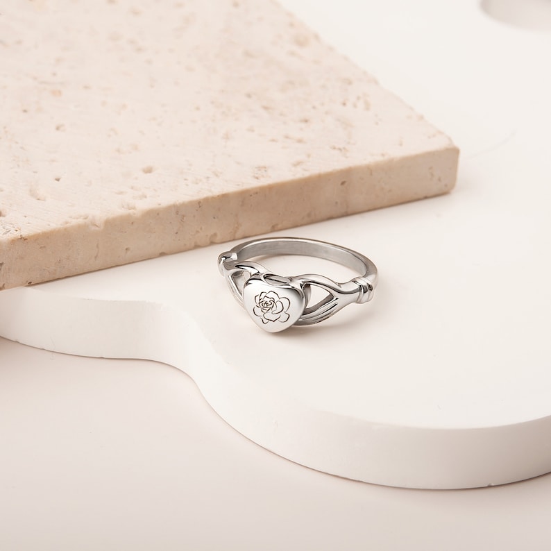 May include: A silver ring with a heart-shaped locket featuring a rose design. The ring is designed to hold a small amount of ashes or other mementos.