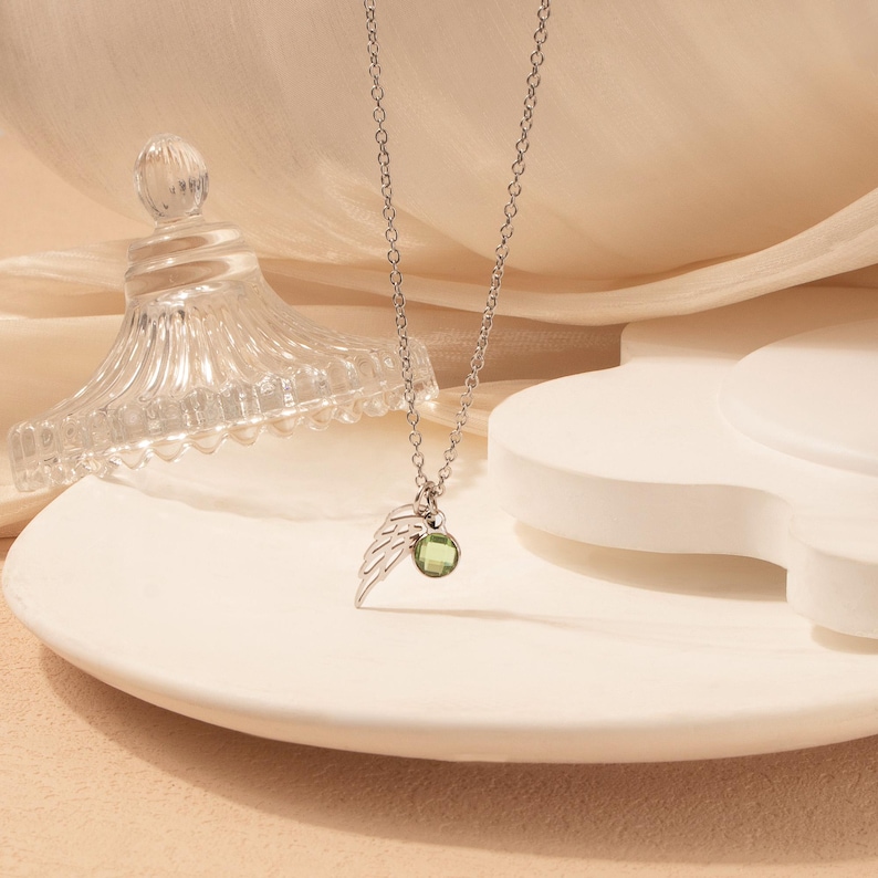 May include: A silver necklace with a delicate chain, featuring a small wing charm and a round, green gemstone pendant. The necklace is displayed on a white, curved surface, with a glass jar in the background.